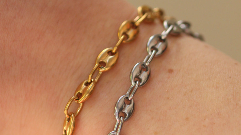 Two metal chain bracelets, one gold-toned and one silver-toned, worn stacked on a person's wrist against a soft beige background.