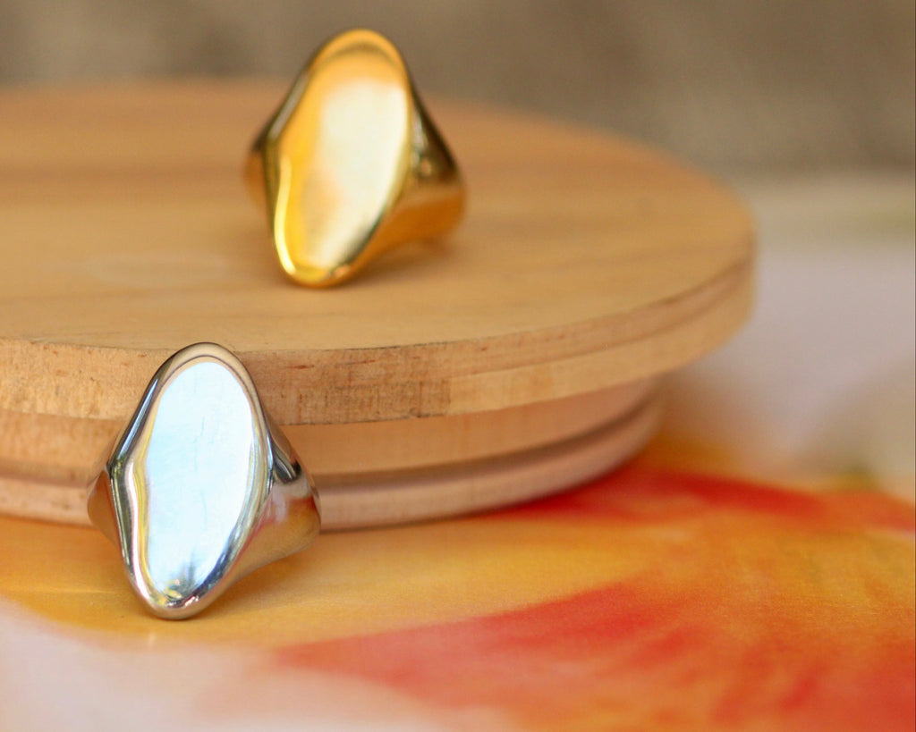 Two minimalist signet-style rings with organic, polished oval-shaped stones, one in polished gold and one in polished silver, displayed on a round wooden riser.