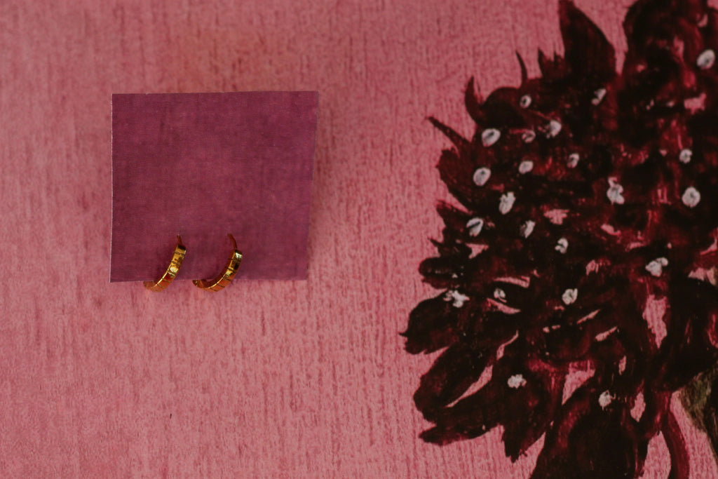 Two small gold rings displayed on a small purple square card against a pink textured background with a dark floral print in the top corner.