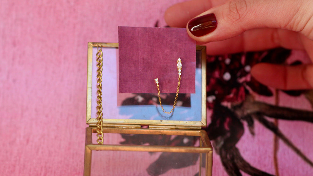 A gold chain earring with a small clear stone is displayed on a purple card inside a gold-framed glass case, held by a hand with dark red nail polish against a pink background.