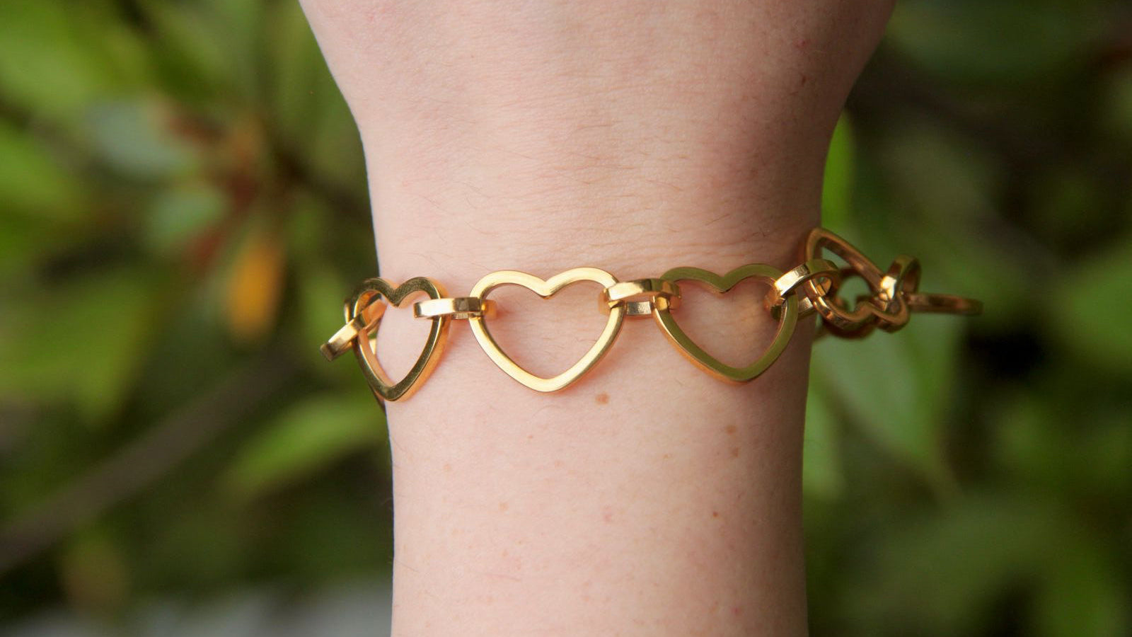 Gold heart-shaped bracelet on a wrist with a blurred green background